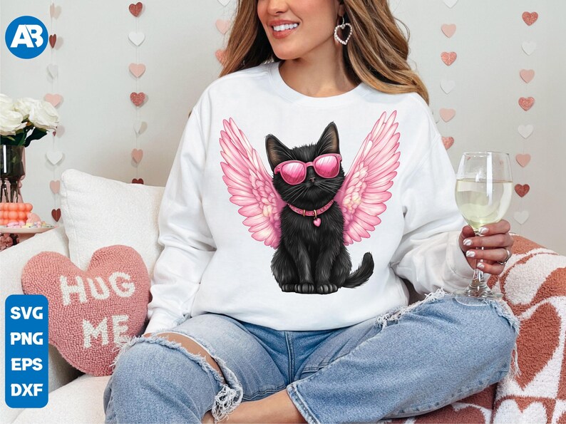 May include: A white sweatshirt with a black cat wearing pink angel wings and sunglasses. The cat is sitting on a white background. The text "Hug Me" is visible on a pink heart-shaped pillow in the background.