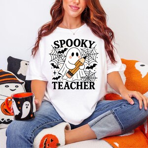 May include: White t-shirt with the text "SPOOKY TEACHER" and a graphic of a ghost holding a pencil, surrounded by bats and spiderwebs. The image also includes a black mug with ghost and pumpkin designs.