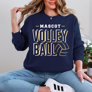 May include: A navy blue sweatshirt with the text "MASCOT VOLLEYBALL" in gold and white lettering. A gold volleyball graphic is to the right of the text.