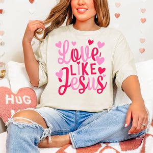 May include: A white t-shirt with a pink and red graphic that says "Love Like Jesus" with pink hearts around the text. The shirt is being worn by a person with long brown hair.