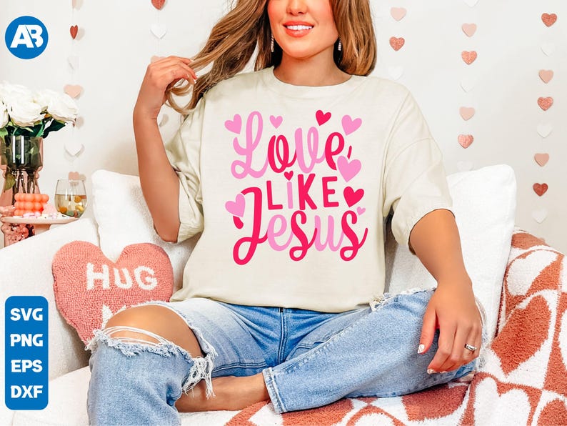 May include: A white t-shirt with a pink and red graphic that says "Love Like Jesus" with pink hearts around the text. The shirt is being worn by a person with long brown hair.