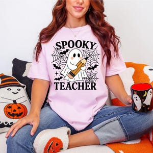 May include: A light pink t-shirt with the words "SPOOKY TEACHER" and a graphic of a ghost holding a pencil, surrounded by bats and spiderwebs. The person is wearing blue jeans and holding a Halloween-themed mug.