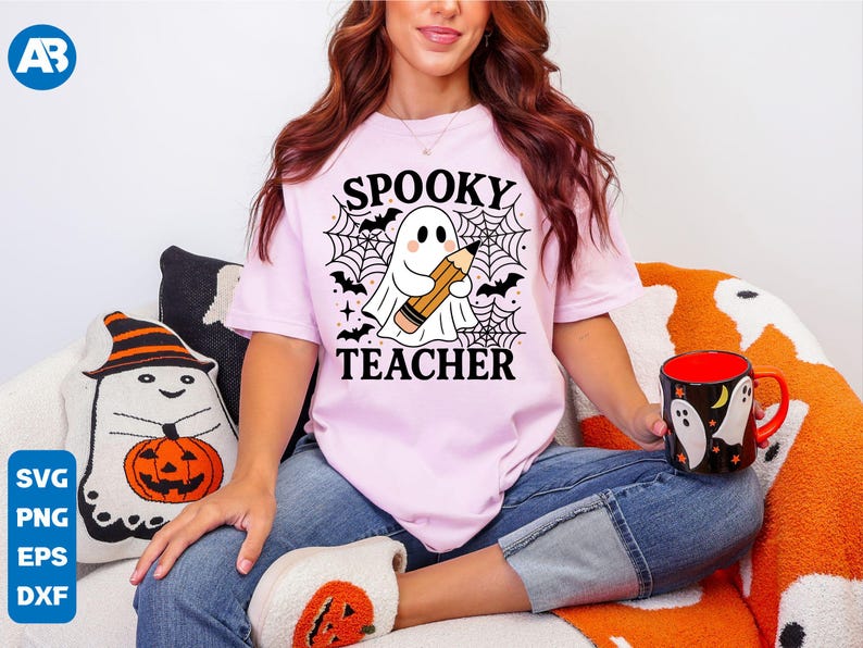 May include: A light pink t-shirt with the words "SPOOKY TEACHER" and a graphic of a ghost holding a pencil, surrounded by bats and spiderwebs. The person is wearing blue jeans and holding a Halloween-themed mug.