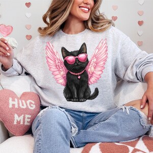 May include: A gray sweatshirt with a black cat wearing pink angel wings and sunglasses. The cat is wearing a pink collar with a heart charm. The text "Hug Me" is on a pink heart-shaped pillow in the background.