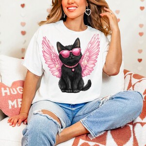 May include: A white t-shirt with a black cat wearing pink angel wings and sunglasses. The cat is sitting on a white background. The t-shirt is being worn by a person.