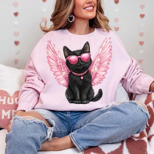 May include: A pink sweatshirt with a black cat wearing pink sunglasses and angel wings. The cat is wearing a black collar with a heart charm.