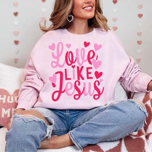 May include: A pink sweatshirt with the text "Love Like Jesus" in pink and red lettering with hearts around the text. The sweatshirt is being worn by a person sitting on a white and brown patterned surface.