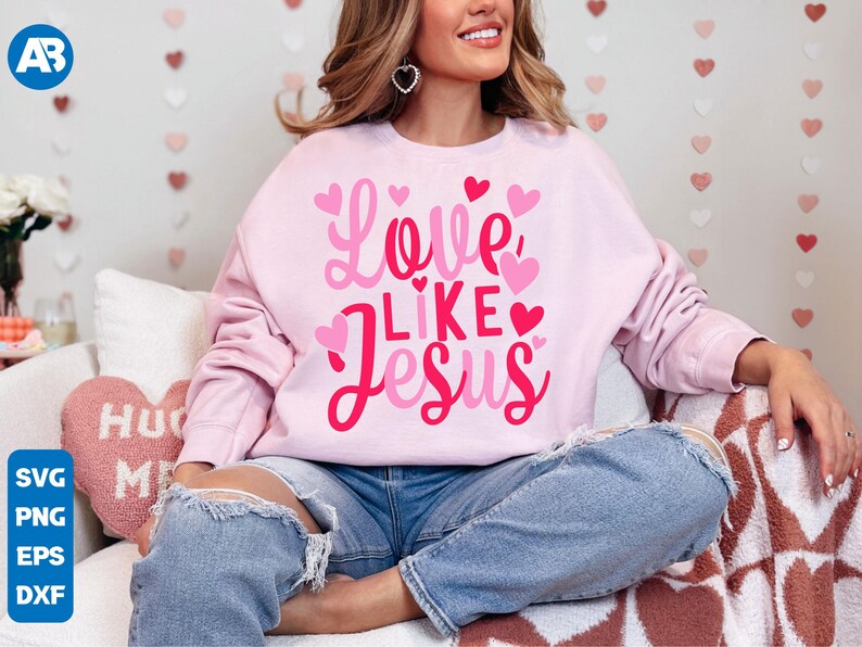 May include: A pink sweatshirt with the text "Love Like Jesus" in pink and red lettering with hearts around the text. The sweatshirt is being worn by a person sitting on a white and brown patterned surface.
