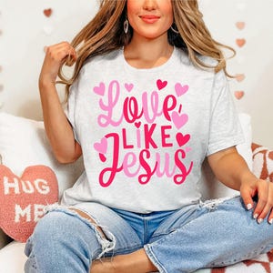 May include: A white t-shirt with a pink graphic that says "Love Like Jesus" surrounded by pink hearts. The shirt is being worn by a person with long brown hair.