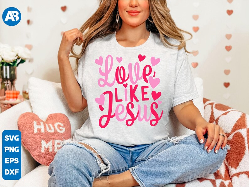 May include: A white t-shirt with a pink graphic that says "Love Like Jesus" surrounded by pink hearts. The shirt is being worn by a person with long brown hair.