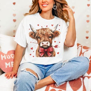 Pu&ograve; includere: Una maglietta bianca con una grafica di una mucca Highland marrone e bianca che indossa un papillon rosso e tiene una freccia rossa in bocca. La mucca &egrave; circondata da cuori rossi.