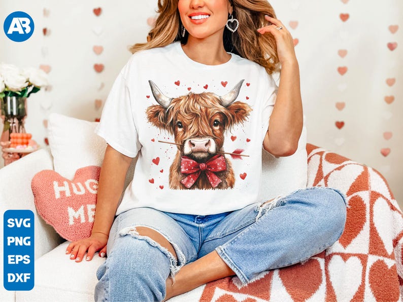 Pu&ograve; includere: Una maglietta bianca con una grafica di una mucca Highland marrone e bianca che indossa un papillon rosso e tiene una freccia rossa in bocca. La mucca &egrave; circondata da cuori rossi.