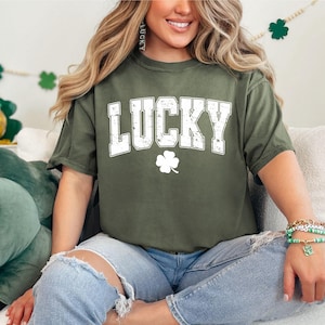 May include: A green t-shirt with the word "LUCKY" in white letters and a white four-leaf clover. The shirt is being worn by a person with long brown hair.