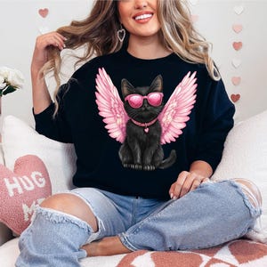 May include: A black sweatshirt with a graphic of a black cat wearing pink angel wings and sunglasses. The cat is sitting on a white background. The sweatshirt is being worn by a person sitting on a couch.
