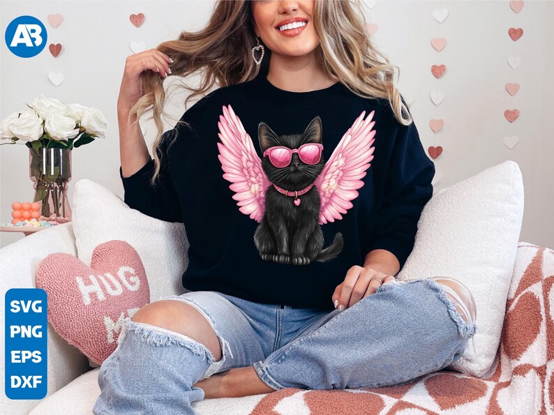 May include: A black sweatshirt with a graphic of a black cat wearing pink angel wings and sunglasses. The cat is sitting on a white background. The sweatshirt is being worn by a person sitting on a couch.