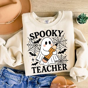 May include: Cream-colored sweatshirt with the words "SPOOKY TEACHER" and a ghost holding a pencil. The design includes bats, spiderwebs, and stars. The sweatshirt is paired with blue jeans, a hat, and sandals.