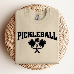 May include: A beige sweatshirt with the text "Pickleball" in black letters and a graphic of two crossed pickleball paddles and a ball.