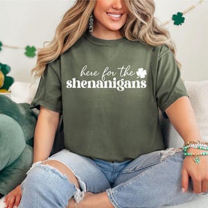May include: A woman wearing a green t-shirt with the text "here for the shenanigans" and a white shamrock. She is wearing blue jeans and has a green clover charm bracelet on her wrist.