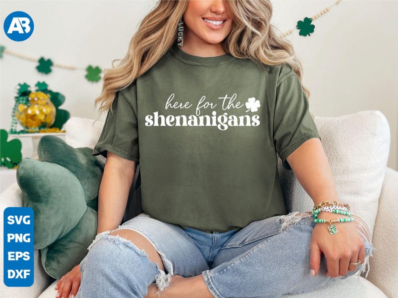 May include: A woman wearing a green t-shirt with the text "here for the shenanigans" and a white shamrock. She is wearing blue jeans and has a green clover charm bracelet on her wrist.