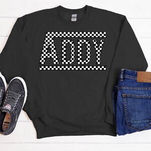 May include: A dark gray sweatshirt with the name "ADDY" printed in white letters on a black and white checkered background.