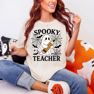 May include: Cream-colored t-shirt with the words "SPOOKY TEACHER" and a ghost holding a pencil, surrounded by bats and spiderwebs. A black and white mug with a ghost design is held in the hand. The theme is Halloween.