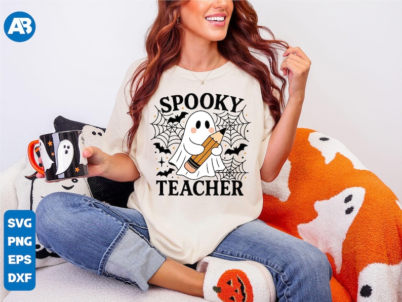 May include: Cream-colored t-shirt with the words "SPOOKY TEACHER" and a ghost holding a pencil, surrounded by bats and spiderwebs. A black and white mug with a ghost design is held in the hand. The theme is Halloween.