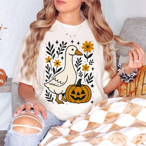 May include: Cream-colored t-shirt featuring a whimsical illustration of a goose, a jack-o'-lantern, and floral elements. The design includes the text "SVG, PNG, EPS, DXF" in a blue box. The overall theme is autumn and Halloween.