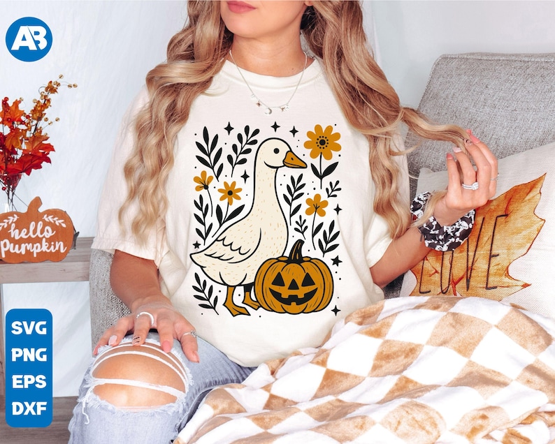 May include: Cream-colored t-shirt featuring a whimsical illustration of a goose, a jack-o'-lantern, and floral elements. The design includes the text "SVG, PNG, EPS, DXF" in a blue box. The overall theme is autumn and Halloween.