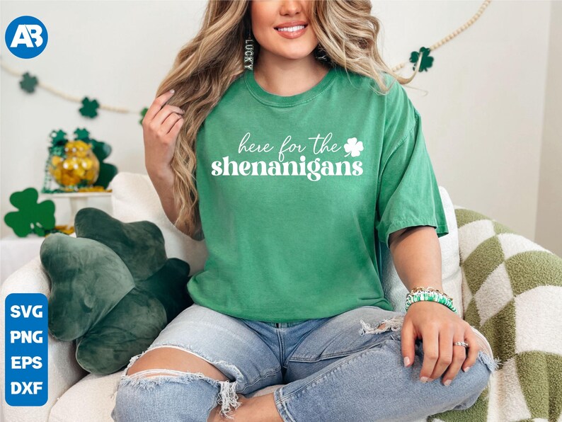 May include: A woman wearing a green t-shirt with the text "here for the shenanigans" and a white shamrock. She is sitting on a couch with a green shamrock pillow.