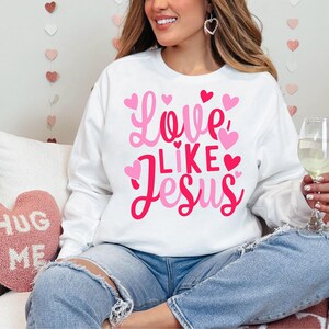 May include: A white sweatshirt with the text "Love Like Jesus" in pink lettering and pink hearts. The sweatshirt is being worn by a person sitting on a couch.