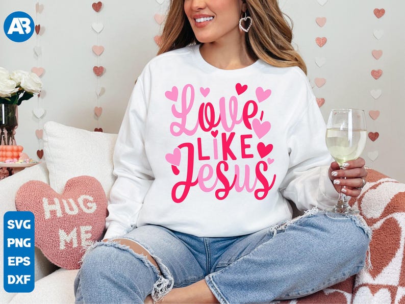 May include: A white sweatshirt with the text "Love Like Jesus" in pink lettering and pink hearts. The sweatshirt is being worn by a person sitting on a couch.