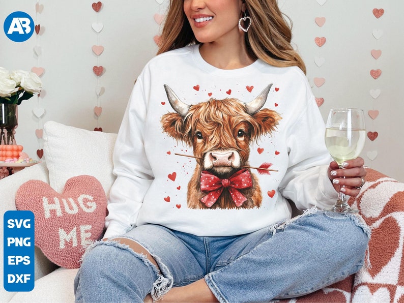 Pu&ograve; includere: Felpa bianca con una mucca delle Highlands marrone e bianca che indossa un fiocco rosso e tiene una freccia, con cuori sullo sfondo.