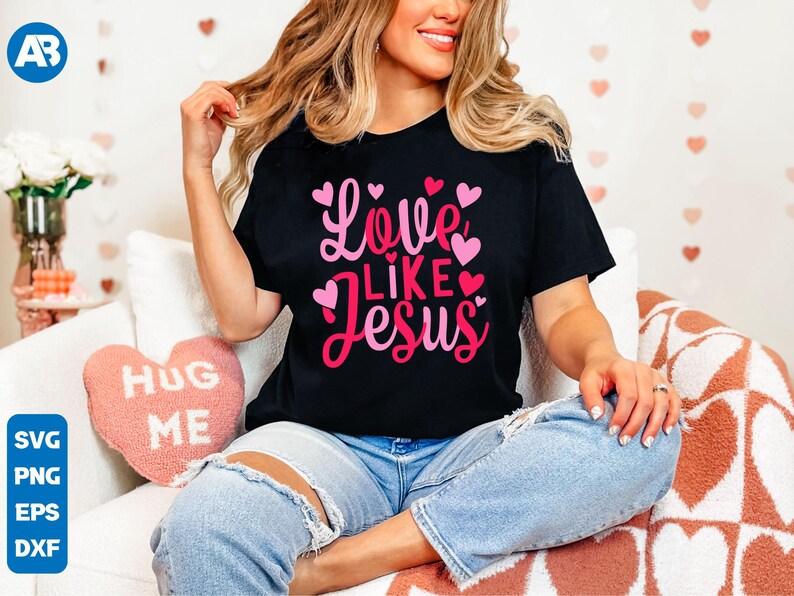 May include: A black t-shirt with the text "Love Like Jesus" in pink and red lettering, surrounded by pink hearts. The shirt is being worn by a person sitting on a pink and white heart-shaped pillow.