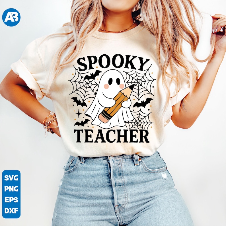May include: Cream-colored t-shirt featuring a Halloween-themed graphic. The design includes a cartoon ghost holding a pencil, surrounded by a spiderweb, bats, and the words "Spooky Teacher". The shirt is paired with blue jeans.
