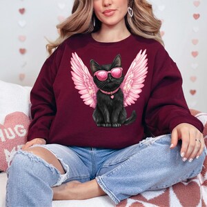 May include: A maroon sweatshirt with a black cat wearing pink angel wings and sunglasses. The cat is wearing a black collar with a silver heart charm.