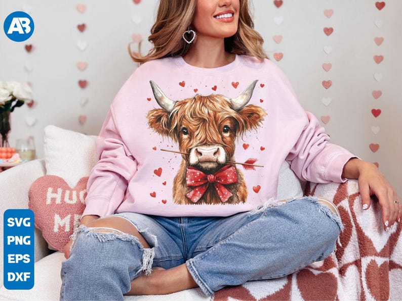Pu&ograve; includere: Una felpa rosa con una simpatica illustrazione di una mucca delle Highlands che indossa un fiocco rosso e tiene una freccia nel naso. La mucca &egrave; circondata da piccoli cuori rosa. La felpa &egrave; indossata da una persona seduta su una coperta a motivi bianchi e rosa.