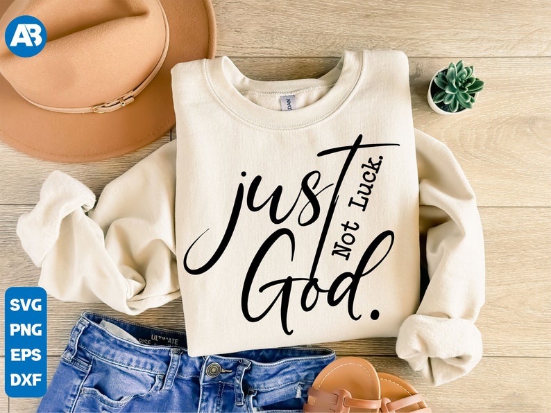 May include: A white sweatshirt with the text "just God. Not luck." in black cursive lettering. The sweatshirt is folded and laid out on a wooden surface with a brown hat, blue jeans, and sandals.