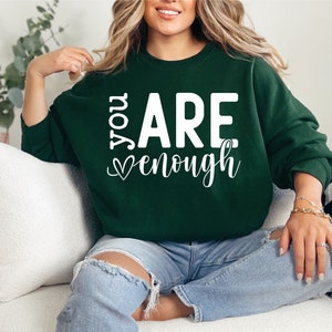May include: A dark green sweatshirt with the text "You Are Enough" in white lettering with a heart symbol. The sweatshirt is being worn by a person sitting on a white couch.
