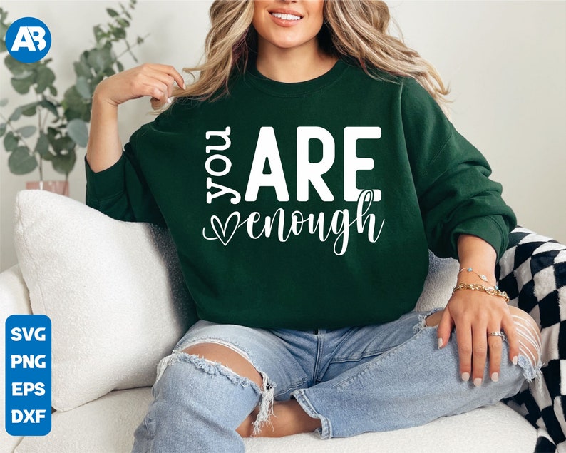 May include: A dark green sweatshirt with the text "You Are Enough" in white lettering with a heart symbol. The sweatshirt is being worn by a person sitting on a white couch.