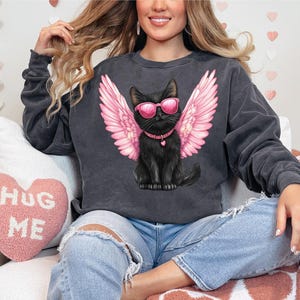 May include: A black sweatshirt with a graphic of a black cat wearing pink sunglasses and pink angel wings. The cat is wearing a black collar with a silver heart charm. The sweatshirt is on a person wearing blue jeans.