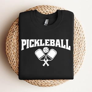 May include: Black t-shirt with white text that says "Pickleball" and a graphic of two crossed pickleball paddles and a ball.