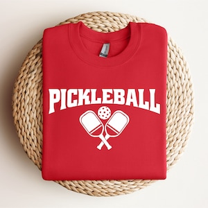 May include: Red t-shirt with white text that says "Pickleball" and a graphic of two crossed pickleball paddles and a ball.