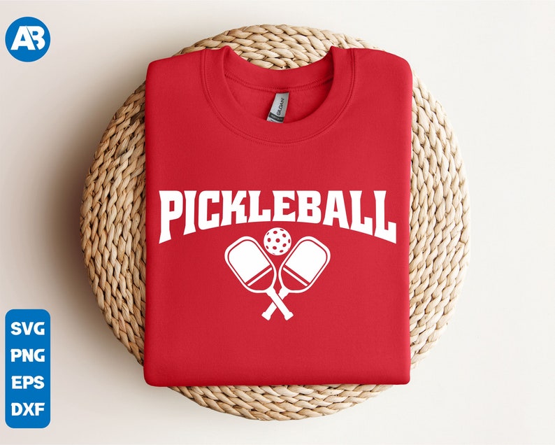 May include: Red t-shirt with white text that says "Pickleball" and a graphic of two crossed pickleball paddles and a ball.