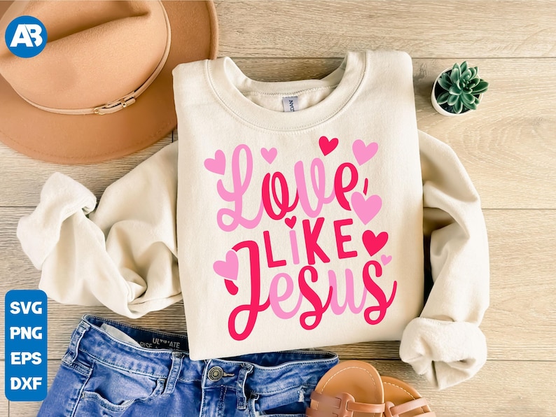 May include: A white sweatshirt with the text "Love Like Jesus" in pink and red lettering. The text is surrounded by pink hearts. The sweatshirt is laid out on a wooden surface with a brown hat, blue jeans, and a small potted plant.