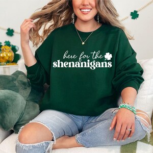 May include: A green sweatshirt with the text "here for the shenanigans" in white lettering and a white shamrock graphic. The model is wearing blue jeans.