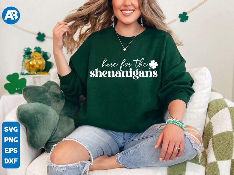 May include: A green sweatshirt with the text "here for the shenanigans" in white lettering and a white shamrock graphic. The model is wearing blue jeans.