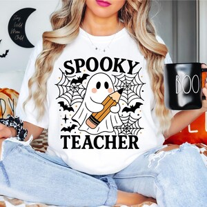 May include: White t-shirt with a Halloween-themed graphic. The design features a ghost holding a pencil, spiderwebs, bats, and the words "Spooky Teacher." A black mug with "BOO" in white is held in the hand.