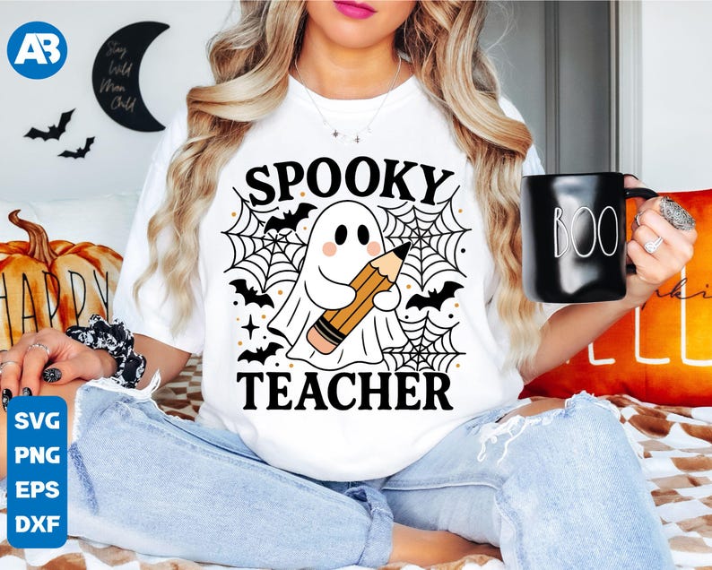 May include: White t-shirt with a Halloween-themed graphic. The design features a ghost holding a pencil, spiderwebs, bats, and the words "Spooky Teacher." A black mug with "BOO" in white is held in the hand.