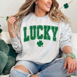 May include: A gray sweatshirt with the word "LUCKY" in green letters with a distressed look. A green four-leaf clover is below the word.