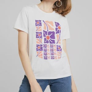 Puede incluir: Una camiseta blanca de manga corta con un estampado gráfico central compuesto por paneles rectangulares. El diseño muestra flores moradas estilizadas con centros naranjas y hojas de color melocotón claro dispuestas en cuadrícula.